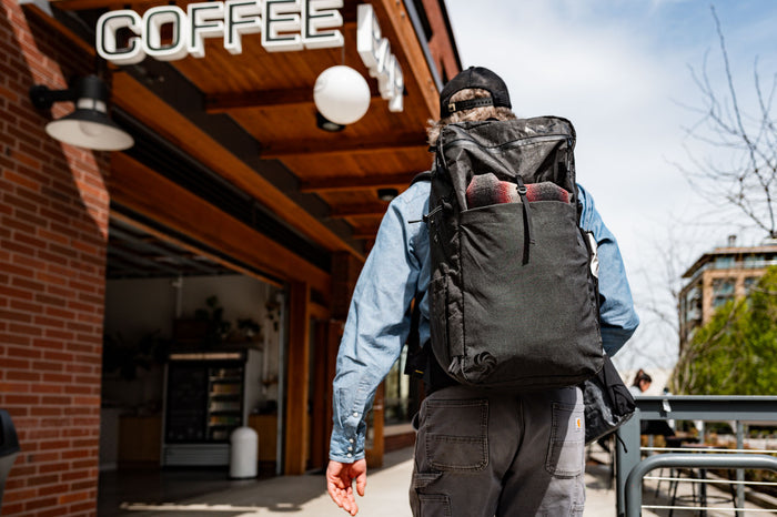 Black ADC pack being worn out in the city with a wool jacket in the rear pocket outside of a coffee shop.