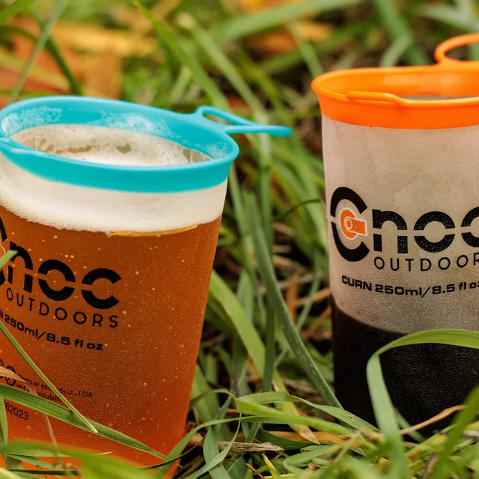 Two Cnoc Outdoors CURN 250ml collapsible cups sitting in green grass, one filled with amber beer topped with foam and fitted with a blue silicone rim, the other half filled with wine with an orange rim.