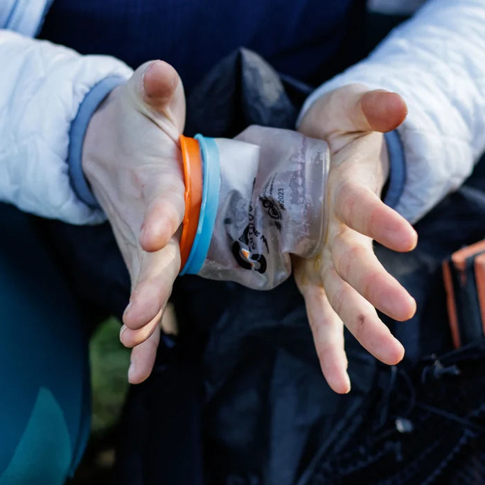 Two hands holding a fully collapsed Cnoc Outdoors CURN cup squeezed flat, with the orange and blue silicone rims stacked together to show its minimal packed profile.