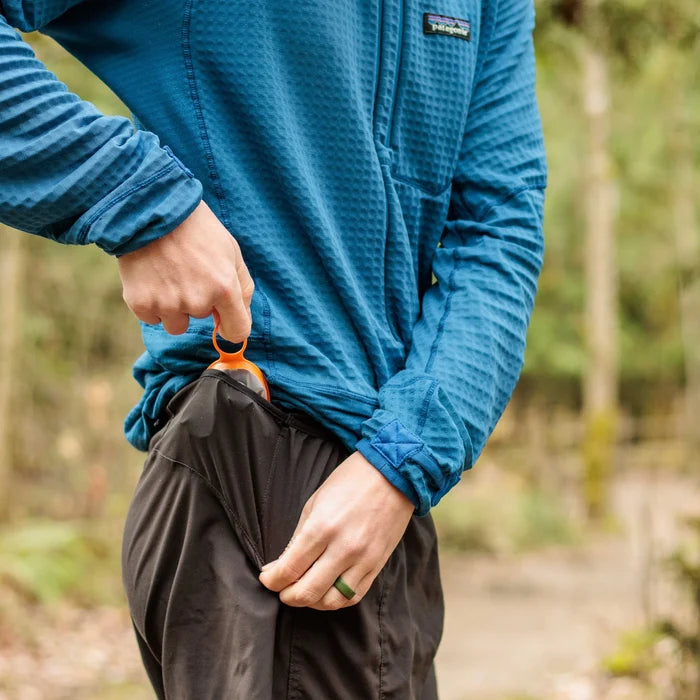 A person on a wooded trail pulling a collapsed Cnoc Outdoors CURN cup with an orange rim from the hip pocket of black shorts, showing its packable size.